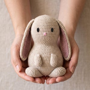 May include: A small, beige crocheted rabbit toy with long, floppy ears featuring a dusty rose pink inner lining. The stuffed animal has two solid black circular eyes and a small, embroidered pink nose, crafted with a consistent yarn stitch.