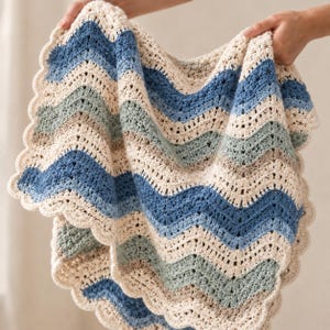 May include: A crocheted blanket featuring a chevron pattern in alternating stripes of cream, sage green, light blue, and dark blue. The edges of the blanket are finished with a decorative scalloped border, creating a textured and soft appearance.