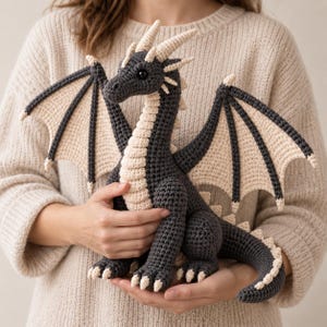 Könnte beinhalten: Ein dunkelgraues und cremefarbenes, gehäkeltes Drachen-Stofftier mit großen Flügeln, einem langen Schwanz und kleinen Hörnern auf dem Kopf. Der Drache hat eine strukturierte Garnoberfläche, schwarze Perlenaugen und cremefarbene Akzente an Bauch und Flügeln.