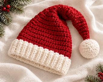 Puede incluir: Un gorro de Papá Noel tejido a ganchillo con una corona puntiaguda de color rojo intenso y un borde grueso y acanalado de color blanco roto. El gorro termina en un pompón redondo y texturizado de color blanco roto unido a la punta del accesorio.
