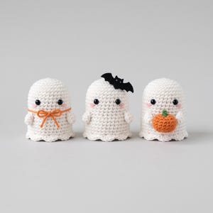 Pode incluir: Três figuras de fantasmas em crochê branco com olhos de contas pretas e bochechas rosadas. O fantasma da esquerda tem um laço laranja, o central tem um morcego de feltro preto na cabeça e o da direita segura uma pequena abóbora de crochê.