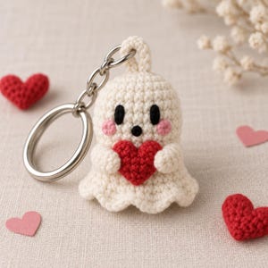 May include: A cream-colored crocheted ghost keychain featuring two black eyes, a small black mouth, and pink cheeks. The ghost is holding a bright red crocheted heart in its arms and is attached to a silver-toned metal split ring keychain.
