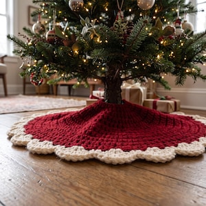 Puede incluir: Una falda de &aacute;rbol de Navidad tejida a ganchillo, redonda, con un centro rojo intenso y un borde festoneado grueso de color crema. El tejido texturizado est&aacute; colocado sobre un suelo de madera, rodeando la base de un &aacute;rbol decorado.