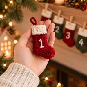 Könnte beinhalten: Ein kleiner, handgehäkelter Weihnachtsstrumpf als Anhänger aus dunkelrotem Garn mit einem cremefarbenen Bündchen und einer passenden Schlaufe zum Aufhängen. Die Zahl Eins ist mit weißem Faden auf die Vorderseite des Strumpfes gestickt.