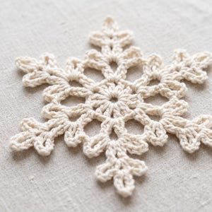 May include: A cream-colored crocheted snowflake ornament featuring a central circular motif with six radiating arms, each ending in a delicate, multi-pointed leaf-like structure created with intricate yarn loops and knots.