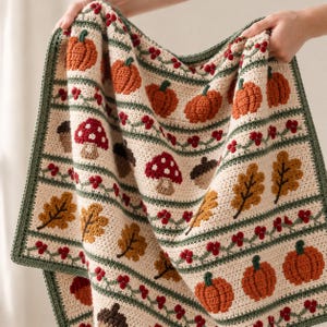 May include: A cream-coloured crochet blanket featuring horizontal bands of autumn-themed motifs including orange pumpkins, red and white mushrooms, brown acorns, and golden oak leaves, separated by thin green stripes with small red berries.