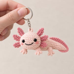 Könnte beinhalten: Ein gehäkelter Axolotl-Schlüsselanhänger mit hellrosa Körper, dunkelrosa Kiemen, einem langen Schwanz und kleinen Gliedmaßen. Die Figur hat zwei schwarze Perlenaugen und einen aufgestickten schwarzen Mund, befestigt an einem silbernen Metallring.