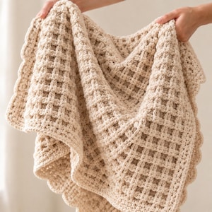 May include: A cream-colored crocheted blanket featuring a prominent waffle stitch texture throughout the main body. The edges of the blanket are finished with a decorative scalloped border, creating a soft and uniform appearance across the fabric.