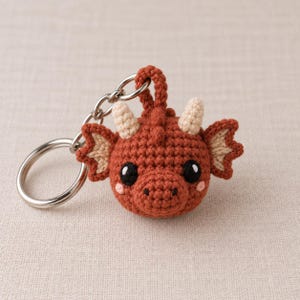 Peut inclure: Un porte-clés dragon en crochet de couleur rouille avec une tête ronde, deux petites cornes crème et des ailes intérieures assorties. Le dragon possède de grands yeux noirs avec des reflets blancs et de petites joues roses brodées.