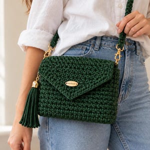 Puede incluir: Un bolso bandolera de ganchillo en color verde oscuro con cierre de solapa, una placa metálica dorada con la inscripción "handmade" y una borla a juego. El bolso cuenta con una correa gruesa y trenzada de color verde oscuro con cierres dorados.