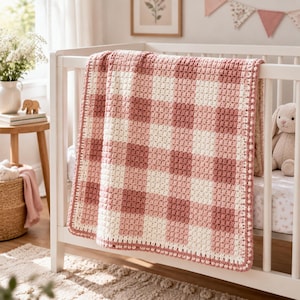 May include: A crochet baby blanket featuring a gingham pattern in shades of dusty rose, light pink, and cream. The blanket is constructed with a textured stitch and finished with a simple border, draped over the side of a white wooden crib.