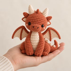 May include: A small, handcrafted crochet dragon toy featuring a rust-orange body with a cream-colored belly, inner wings, and horns. The creature has two black safety eyes, small pink blush marks on its cheeks, and a textured, stitched surface.