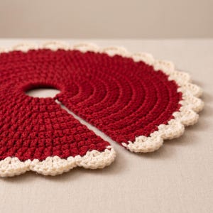 Puede incluir: Una falda de &aacute;rbol circular tejida a ganchillo en color rojo oscuro, con una abertura central y una ranura para facilitar su colocaci&oacute;n. El borde exterior presenta un acabado festoneado en color crema que contrasta con el hilo rojo.