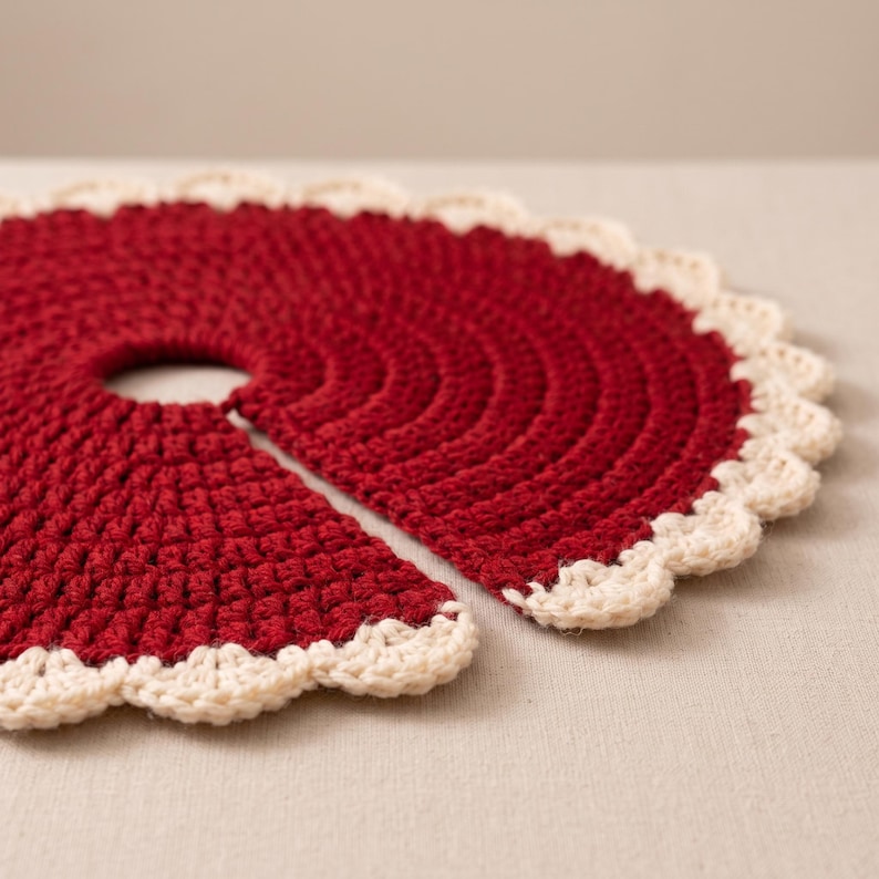 Puede incluir: Una falda de &aacute;rbol circular tejida a ganchillo en color rojo oscuro, con una abertura central y una ranura para facilitar su colocaci&oacute;n. El borde exterior presenta un acabado festoneado en color crema que contrasta con el hilo rojo.