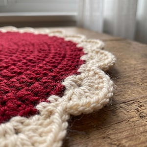 Puede incluir: Un tapete de ganchillo circular que presenta un centro de color rojo intenso hecho con un patr&oacute;n de puntos texturizados. El borde est&aacute; rematado con un fest&oacute;n decorativo de color crema que crea un contorno suave y redondeado.