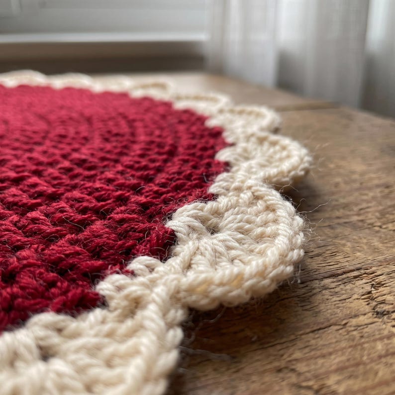 Puede incluir: Un tapete de ganchillo circular que presenta un centro de color rojo intenso hecho con un patr&oacute;n de puntos texturizados. El borde est&aacute; rematado con un fest&oacute;n decorativo de color crema que crea un contorno suave y redondeado.