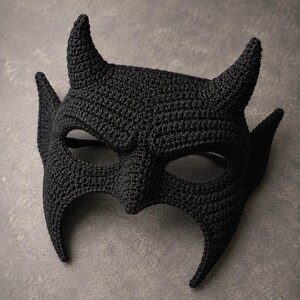 May include: A black crochet mask featuring two pointed horns on the top and two elongated, pointed ear-like structures on the sides. The mask has two large eye openings and a central nose bridge, constructed with a dense, textured yarn stitch pattern.