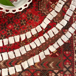 May include: A decorative garland featuring three parallel strands of white fabric squares. Each square is attached to a thin, light-colored string, creating a repeating flag-like pattern with raw, frayed edges along the bottom of each fabric piece.