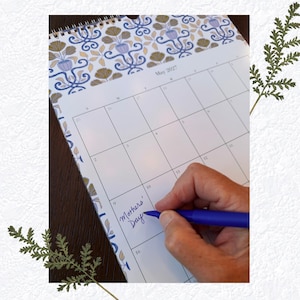 May include: A wall calendar for May 2027 featuring a blue, gold, and white floral pattern at the top. A blue pen is writing the words Mothers' Day in the square for the ninth day of the month. The calendar has a spiral binding at the top edge.