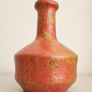 Może przedstawiać: Ceramiczny wazon z szeroką, zaokrągloną podstawą i wąską szyjką. Powierzchnia ma teksturowane, nakrapiane wykończenie w odcieniach żywego koralowego pomarańczu i przygaszonej oliwkowej zieleni, tworząc wyrazisty, nierówny wygląd na całości.