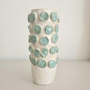 Könnte beinhalten: Eine zylindrische Keramikvase mit einer glänzenden, cremeweißen Grundglasur. Die Außenseite ist mit zahlreichen erhabenen, kreisförmigen, hellblau-grünen Tontellern verziert, die eine gesprenkelte Textur und ein unregelmäßiges Aussehen aufweisen.