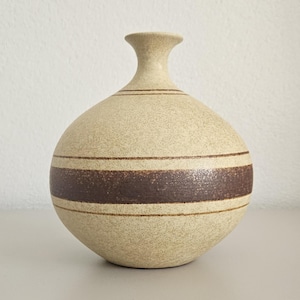 May include: A round ceramic vase with a speckled cream-coloured finish. The vase features a narrow neck and a wide, bulbous body decorated with a prominent horizontal dark brown band flanked by two thinner brown lines encircling the centre.