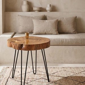 May include: A round side table featuring a natural wood slab top with visible grain and bark edges, supported by three thin black metal hairpin legs. A small, textured gold-toned glass vessel sits on the surface of the wooden tabletop.