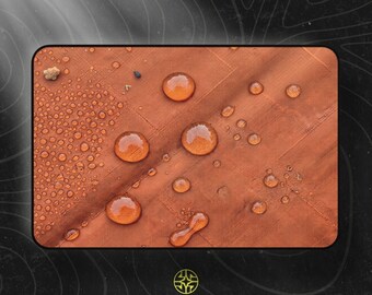 Puede incluir: Una pieza rectangular de tela color óxido con una textura de rejilla, que presenta numerosas gotas de agua de diversos tamaños sobre la superficie. Debajo de la tela, el texto ASSEMBLE WONDER está impreso en blanco con un logotipo circular encima.