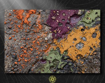 Puede incluir: Una superficie de asfalto texturizada cubierta con parches dispersos de polvo de pigmento naranja vibrante, morado intenso, verde oliva y amarillo mostaza. El suelo es irregular con pequeñas piedras, grietas y zonas húmedas, creando un patrón.
