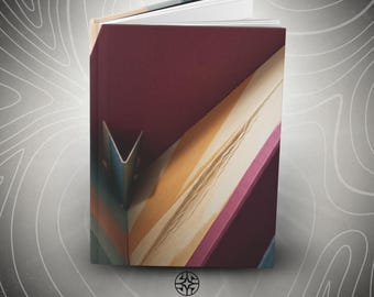 Puede incluir: Un libro de tapa dura con cubierta color granate que presenta un diseño geométrico de rayas diagonales en tonos amarillo, rosa y azul. El libro está ligeramente abierto, mostrando páginas blancas. Debajo, el texto ASSEMBLE WONDER está impreso en negro.