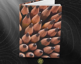 Puede incluir: Un cuaderno de tapa dura con una impresión fotográfica de alta resolución de numerosos lápices de madera afilados con puntas de grafito oscuro. La cubierta muestra un patrón denso de puntas de lápiz en tonos bronceado y marrón. El texto ASSEMBLE WONDER aparece debajo.