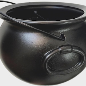 May include: A matte black cauldron with a rounded body, a wide open top, and a thin metal handle attached to the sides. The exterior features a textured surface with a prominent oval-shaped decorative element on the side, providing a sturdy appearance.