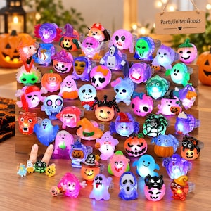 May include: A collection of assorted light-up Halloween rings featuring various designs including pumpkins, ghosts, skulls, spiders, and bats. Each plastic ring glows with internal LED lights, displayed on a tiered wooden stand with a sign.