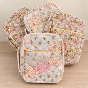 May include: A collection of four quilted fabric cases featuring a floral patchwork pattern in shades of pink, blue, and white. Each case has a gold-toned zipper closure and a handle. The front-most case displays a pink embroidered monogram.