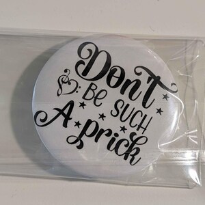 May include: A circular white pin-back button encased in a clear plastic sleeve. The button features black decorative lettering that reads "Don't Be SUCH A prick" with a treble clef symbol replacing the letter o and small stars scattered throughout.