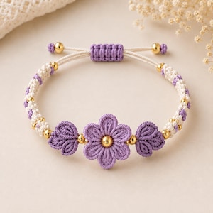 Pode incluir: Uma pulseira de macramé com um motivo central de flor roxa com uma conta dourada no centro, ladeada por dois pequenos detalhes roxos em forma de folha. A banda consiste em cordão atado em creme e roxo com pequenas contas douradas.