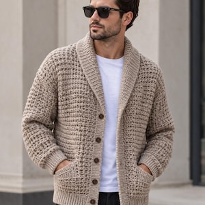 May include: A beige, textured knit cardigan featuring a shawl collar, long sleeves with ribbed cuffs, a ribbed hem, and five brown buttons down the front. The garment includes two front pockets and is worn over a plain white t-shirt and dark pants.