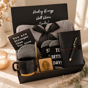 May include: A black gift box containing a grey fuzzy blanket tied with a black ribbon, a black notebook with a gold pen, a black ribbed mug, a grey coaster, and socks with text. A card reads You are Stronger than You know and Healing Energy Well Wishes Smiles.