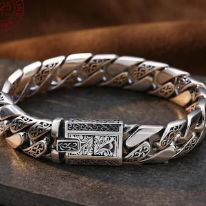 May include: A heavy 925 sterling silver curb chain bracelet featuring polished links with intricate, dark-oxidized floral scrollwork engravings. The bracelet includes a rectangular box clasp mechanism that is also decorated with detailed floral patterns.