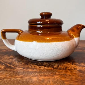 May include: A ceramic teapot featuring a two-tone design with a glossy dark brown lid and upper body, transitioning into a speckled off-white base. The teapot includes a curved handle, a short spout, and a matching removable lid with a knob handle.