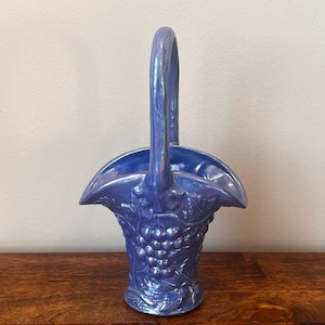 May include: A glossy, periwinkle blue ceramic basket featuring a raised, molded relief pattern of grape clusters and vine leaves on the exterior. The basket has a single, tall, arched handle and a flared, scalloped rim with a smooth, iridescent finish.