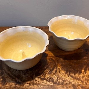 May include: Two ceramic bowls with a pale yellow glaze and a scalloped, wavy rim. Each bowl features a small, pointed protrusion rising from the center of the interior base. The bowls have a smooth, glossy finish and a rounded, deep shape.