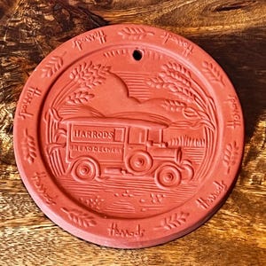May include: A round, terracotta-colored ceramic plaque featuring an embossed design of a vintage delivery truck with the text HARRODS BREAD DELIVERY. The border includes wheat stalks and the repeated word Harrods, with a small hole at the top.