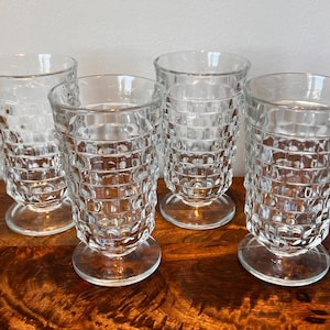 May include: A set of four clear glass tumblers, each featuring a raised grid pattern on the exterior surface. These drinking glasses are designed with a short, circular pedestal base and a slightly flared rim, providing a stable structure.