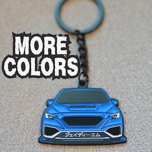 May include: A blue car-shaped keychain with a black metal ring and chain. The front of the car features a black grille, headlights, and a white license plate with Japanese text. The text "MORE COLORS" is printed in white on the upper left side.