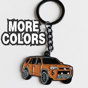 Pode incluir: Um porta-chaves de metal com um pendente em forma de SUV laranja, detalhes pretos e o logótipo da Toyota na grelha frontal. O porta-chaves inclui um anel e uma corrente pretos. O texto MORE COLORS está impresso em branco com um contorno preto.