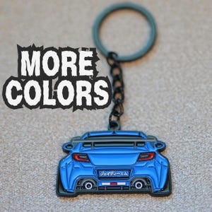 Puede incluir: Un llavero de coche deportivo azul que muestra la parte trasera de un vehículo con alerón, doble tubo de escape y una matrícula con texto en japonés. El llavero incluye una cadena de metal negro y una anilla. El texto MORE COLORS es visible.