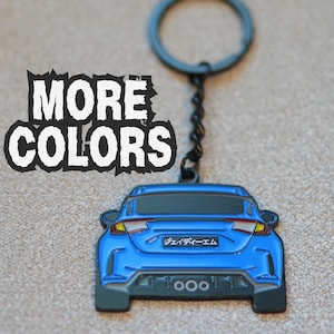 Peut inclure: Un porte-clés en forme de voiture bleue avec un anneau et une chaîne en métal noir. Le design de la voiture montre l'arrière avec des feux rouges et jaunes, une vitre arrière sombre et une plaque avec le texte japonais "ジェイディーエム". Le texte "MORE COLORS" apparaît en blanc avec un contour noir dentelé.