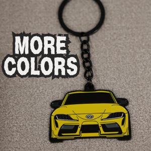 May include: A yellow Toyota Supra car keychain with a black metal chain and ring. The keychain features a detailed front view of the vehicle with black accents and the Toyota logo. The text MORE COLORS is printed in white with a jagged black outline.