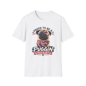 May include: A white short-sleeve t-shirt featuring a graphic of a pug wearing sunglasses with an American flag pattern and a matching flag-patterned bandana. The text reads PROUD TO BE A PUGGIN PATRIOT with small paw prints and stars surrounding the design.